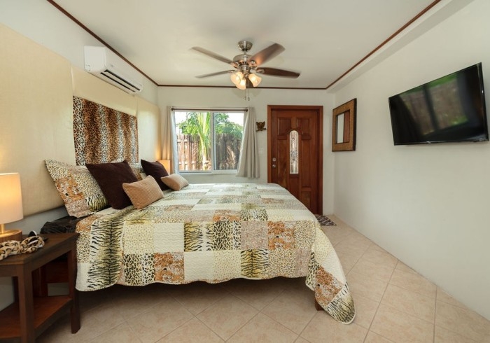 Bed with high upholstered headboard with animal print throw pillows and quilt in neutral colors, nightstands with lamps, ceiling fan, TV on wall, window, and exterior door