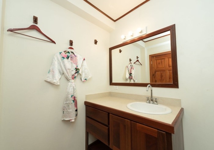 Wood vanity with white counter and sink, mirror with lights, and floral robe hanging on wall