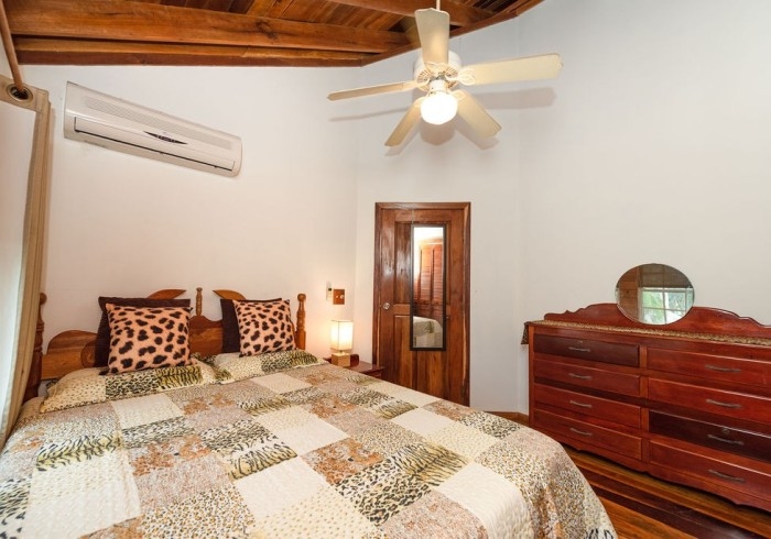 Bedroom with white walls, bed with wooden headboard and quilt and decorative pillows in neutral animal prints, wide wooden dresser with mirror, wooden vaulted ceiling with fan