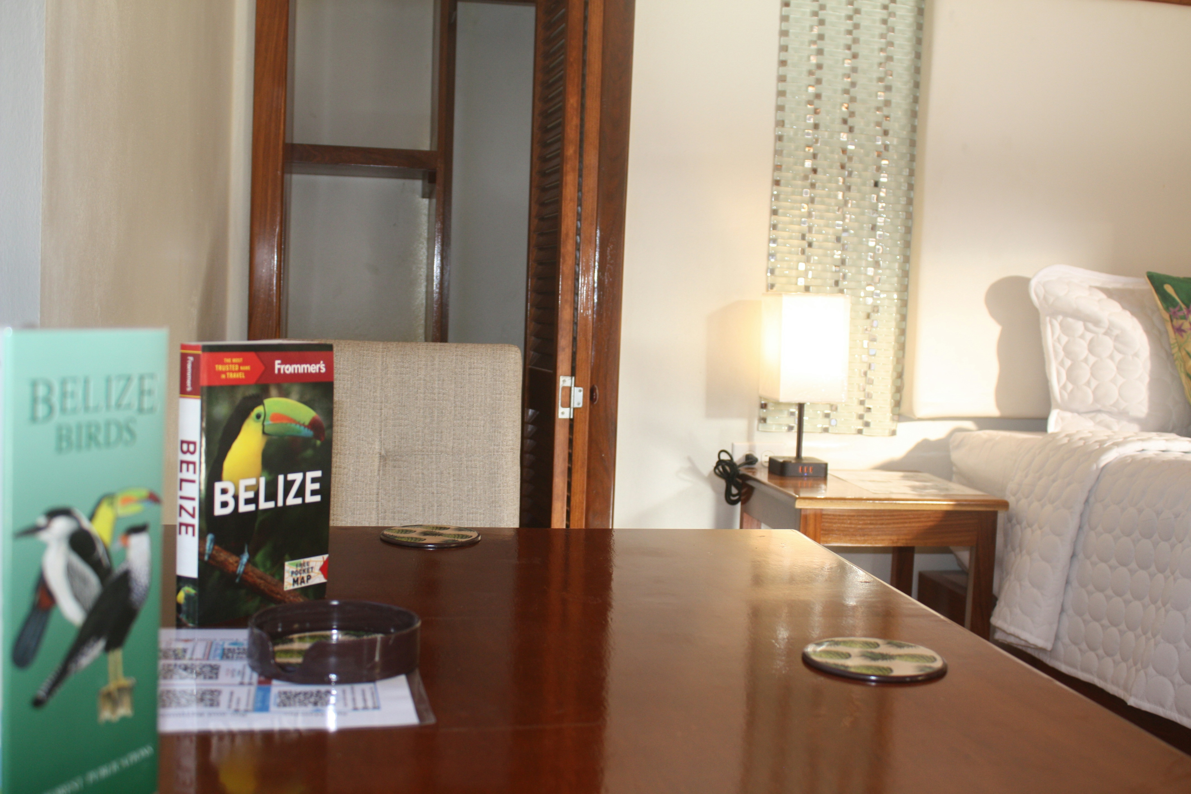 Wood table in bedroom with Belize travel books on top, nightstand with lamp and corner of bed in background