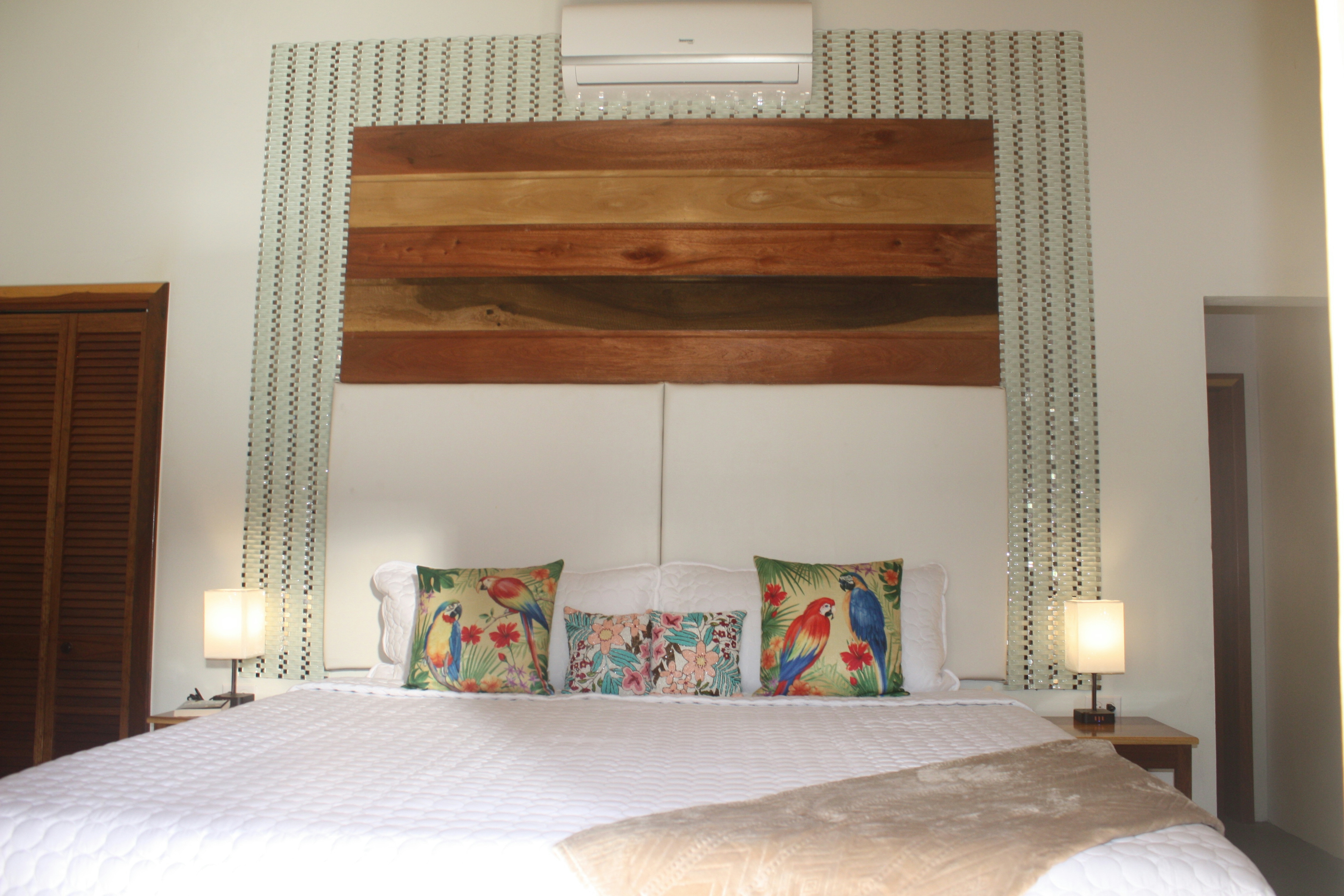 View from foot of bed toward high wooden and upholstered headboard, white quilted bedcover, decorative throw pillows with parrots, nightstands with matching lamps on each side of bed