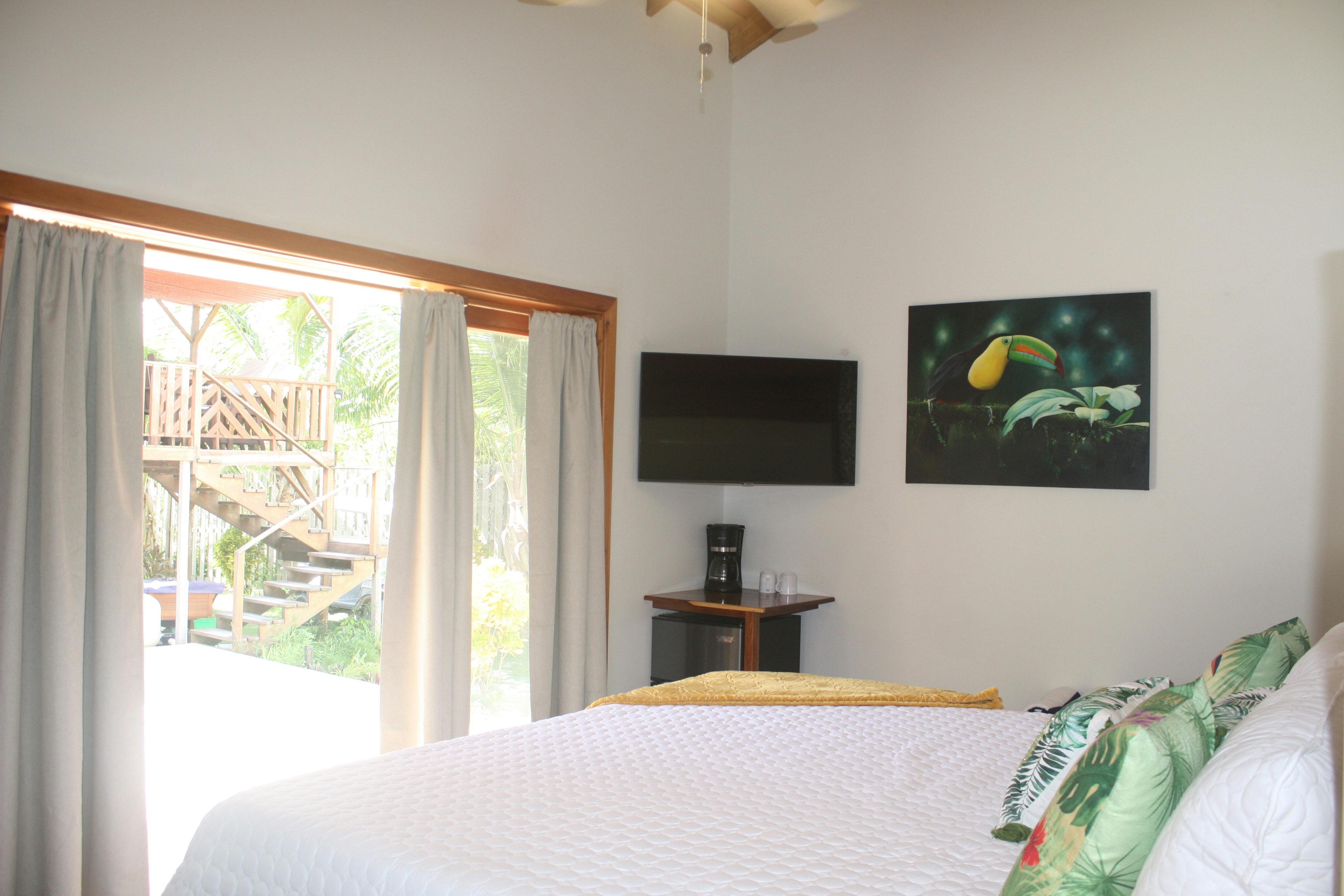 Bed with white cover and pillows with tropical foliage, colorful toucan painting on wall, TV, mini fridge, french doors leading outside