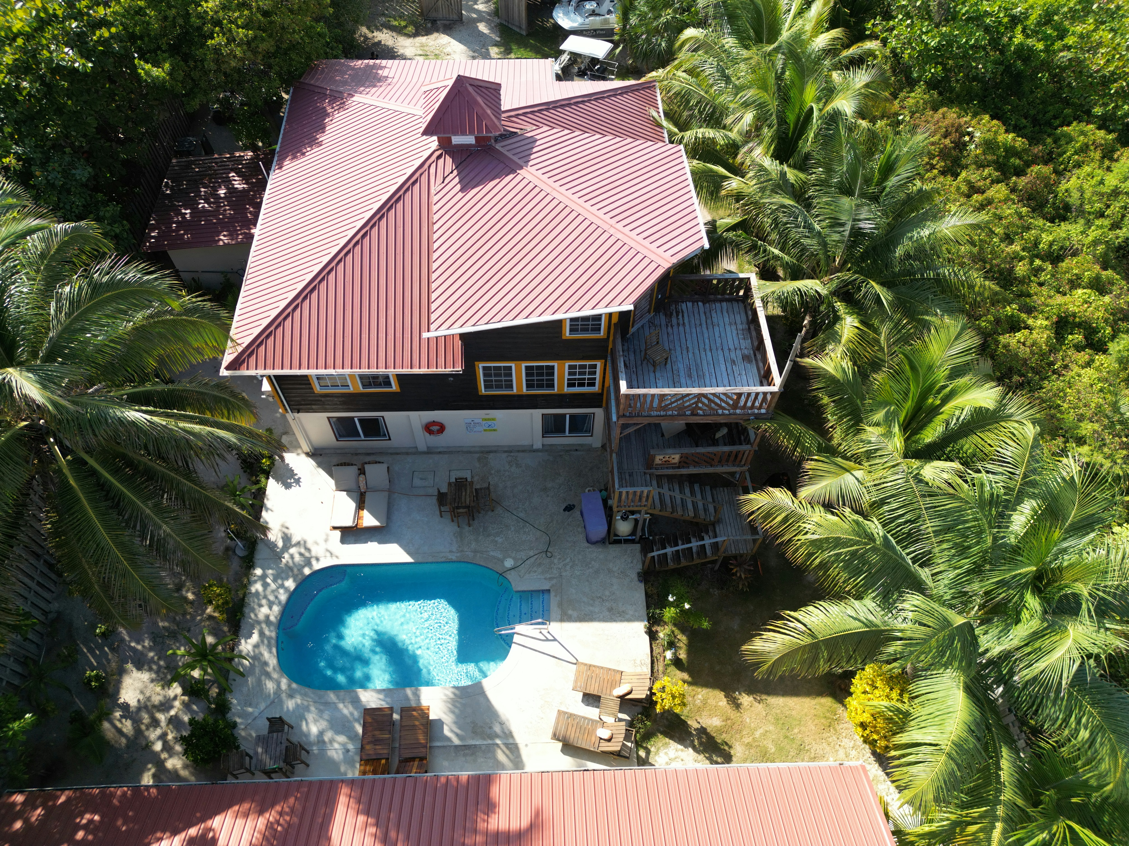 View from above wood three story building with red roof, exterior wood stairs and balconies, pool area with lounge chairs, surrounded by palm trees