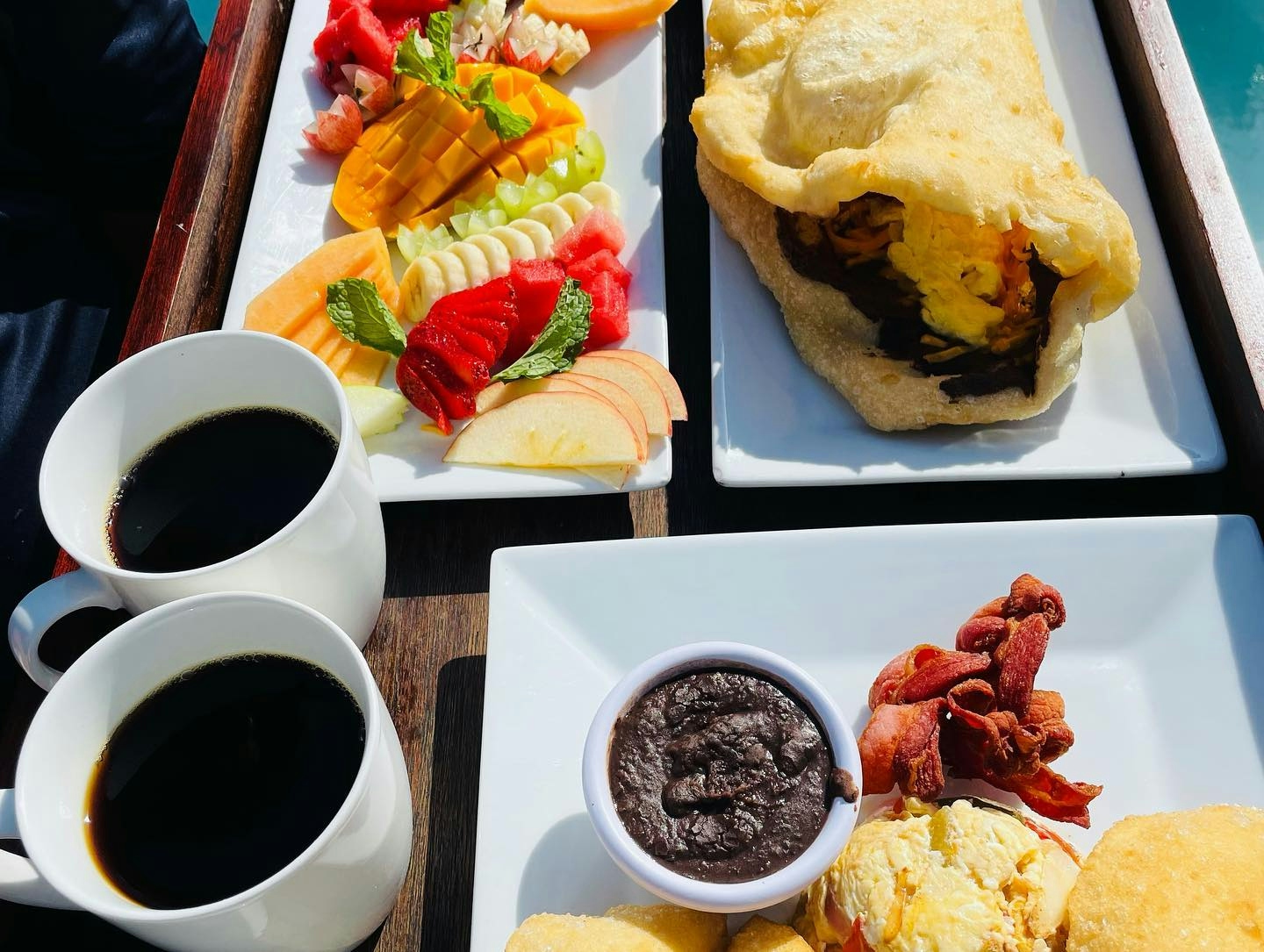 Table set with two mugs filled with coffee and white plates filled with fresh sliced fruit, a breakfast burrito, bacon, and chocolate cake