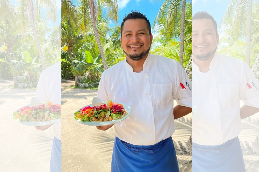 Smiling man in a white chef’s jacket and blue apron presenting a white platter filled with tacos topped with colorful vegetables