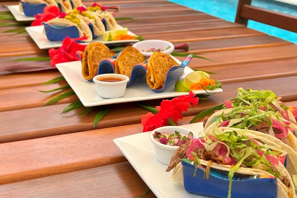 An outdoor table set with square white platters holding tacos made with various toppings and ramekins with sauces on the side