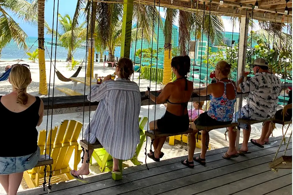 People with backs to camera sitting on swings at bar looking toward sandy beach with hammocks and palm trees and ocean beyond