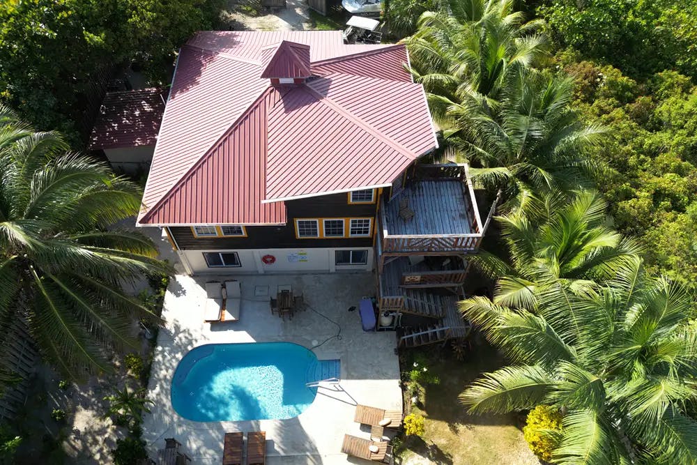 View from above wood three story building with red roof, exterior wood stairs and balconies, pool area with lounge chairs, surrounded by palm trees