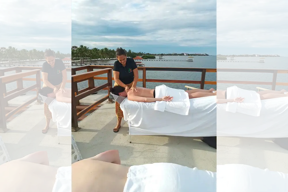 Masseuse rubbing back and shoulders of person lying face down on massage table draped in white towels on deck overlooking ocean