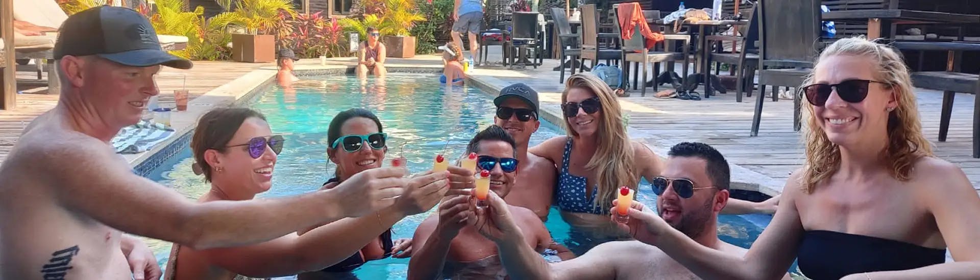 Group of smiling adults in swimsuits at end of pool toasting each other with tropical drinks