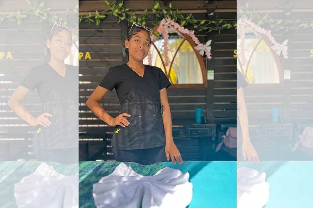 Smiling woman in spa uniform posing behind table with white towel folded like a fan and tropical flower garland behind her