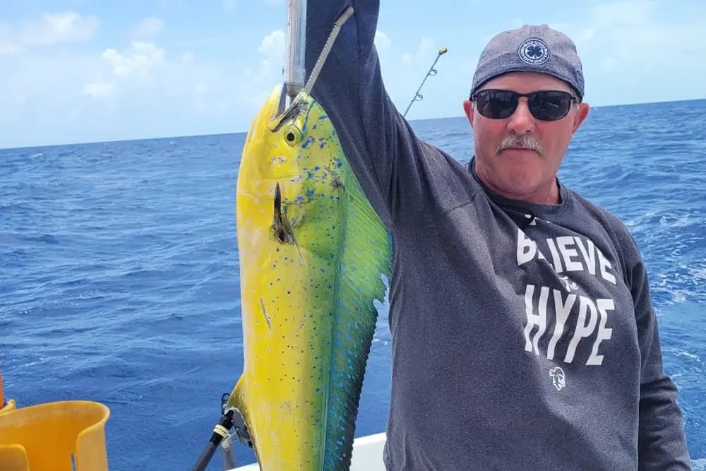 Man with sunglasses on boat deck and holding up fishing line with large yellow fish with blue speckles, ocean waters and blue sky in background