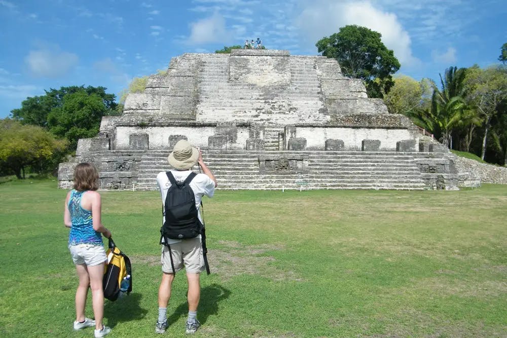 Two adults standing on green grass in front of ancient pyramid ruin with children posing on top