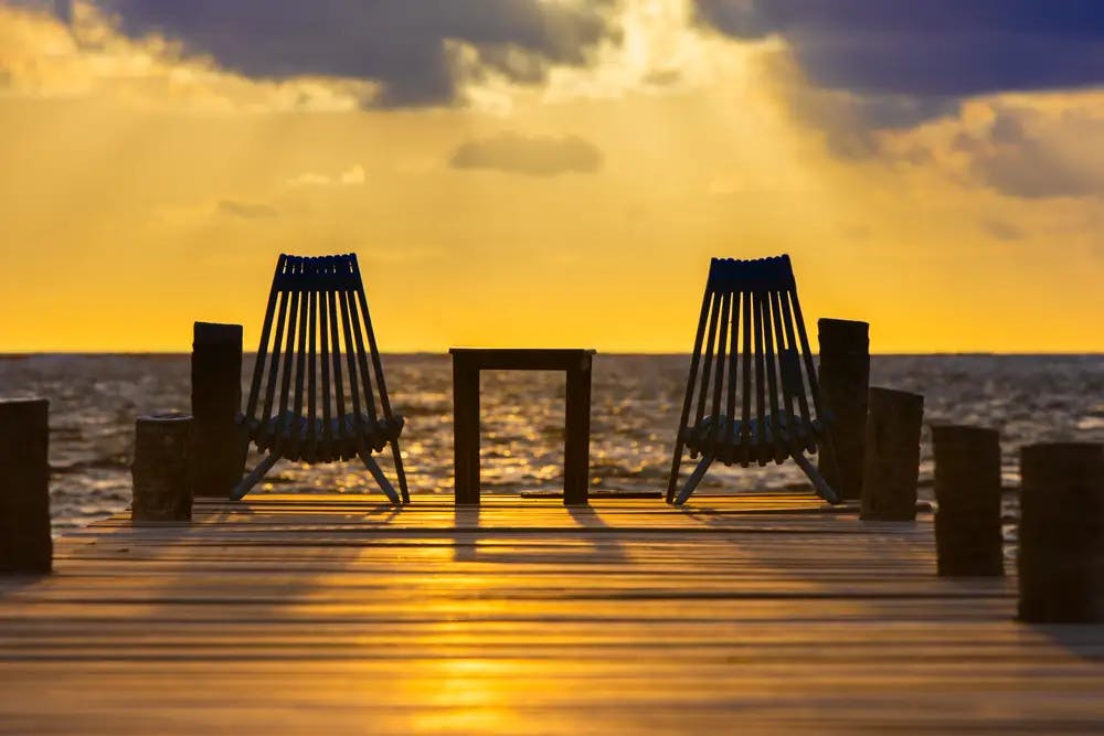 Two wooden chairs silhouetted at end of dock facing sunset over the ocean