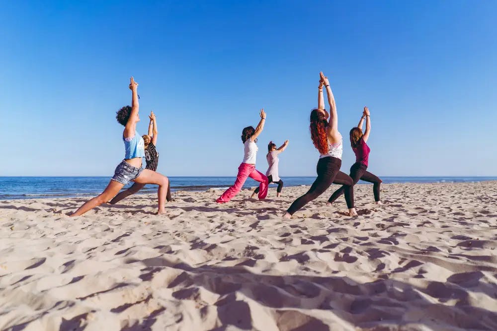 Group of people in fitness clothing doing yoga poses on sandy beach with ocean in background and brilliant cloudless blue sky