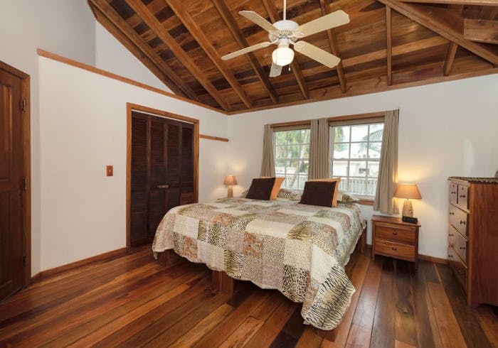 Bedroom with white walls, finished wood floors, and vaulted wood ceilings, ceiling fan, bed with quilt featuring animal print designs, wood dresser, nightstand with lamp, and closet door