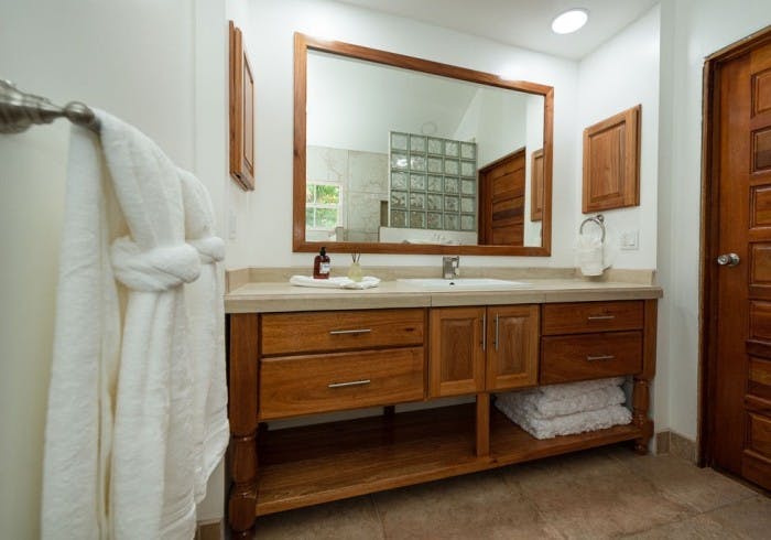 Wooden bathroom vanity with sink, fluffy white towels on rack and shelves, long mirror showing reflection of tiled shower