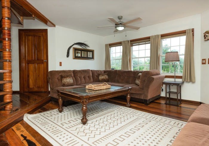 Living room with plush taupe sofa, wooden coffee table, end table with lamp, windows with taupe curtains, ceiling fan, finished wooden floor with neutral woven area rug