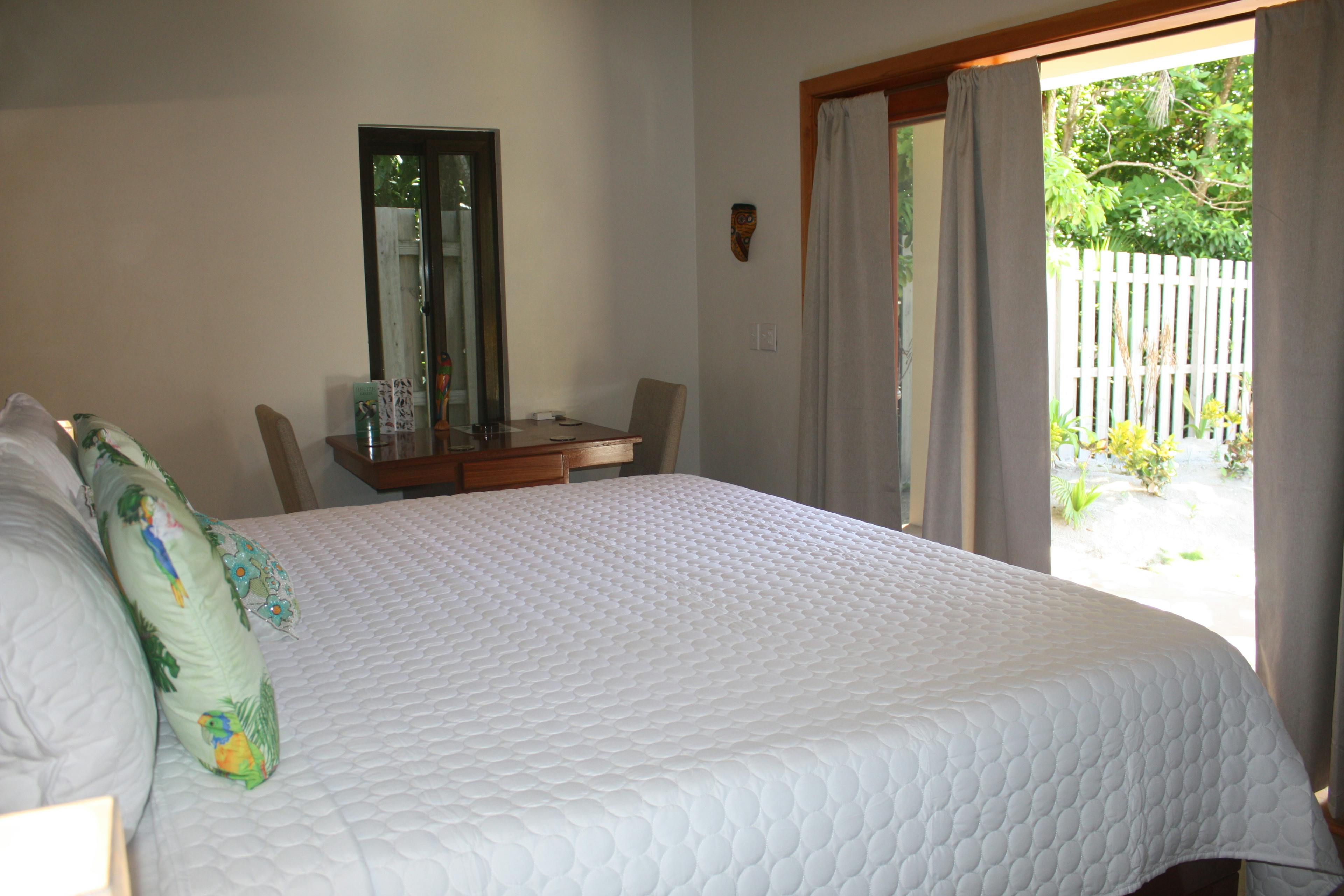 Bed with white quilted bed cover and floral throw pillows, small wooden table with two chairs against the far wall, and french doors with drapes leading to pool deck