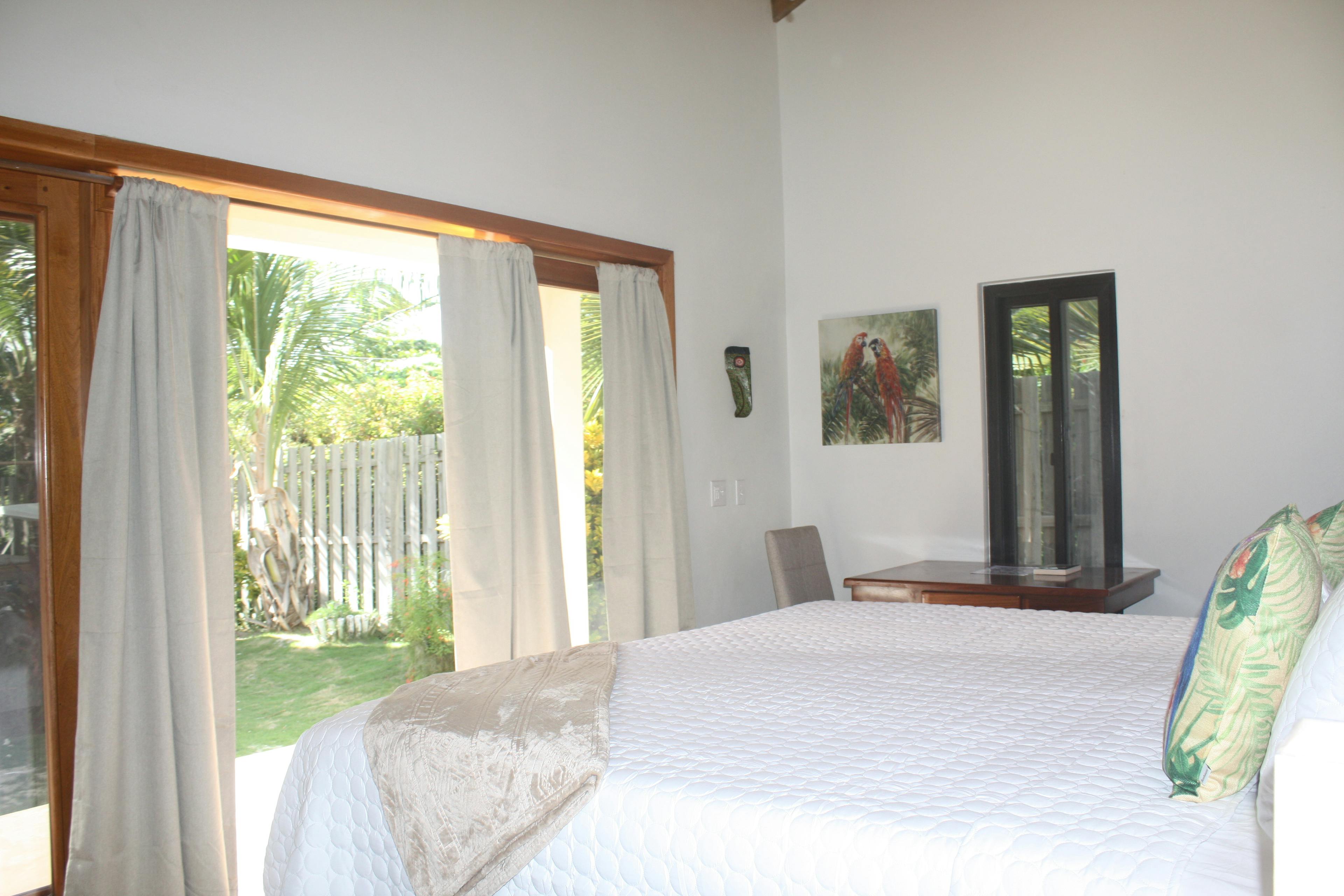 Bed with white quilted bedcover and tropical floral throw pillows, small table with chair and picture of two red macaws hanging on wall, french doors with drapes