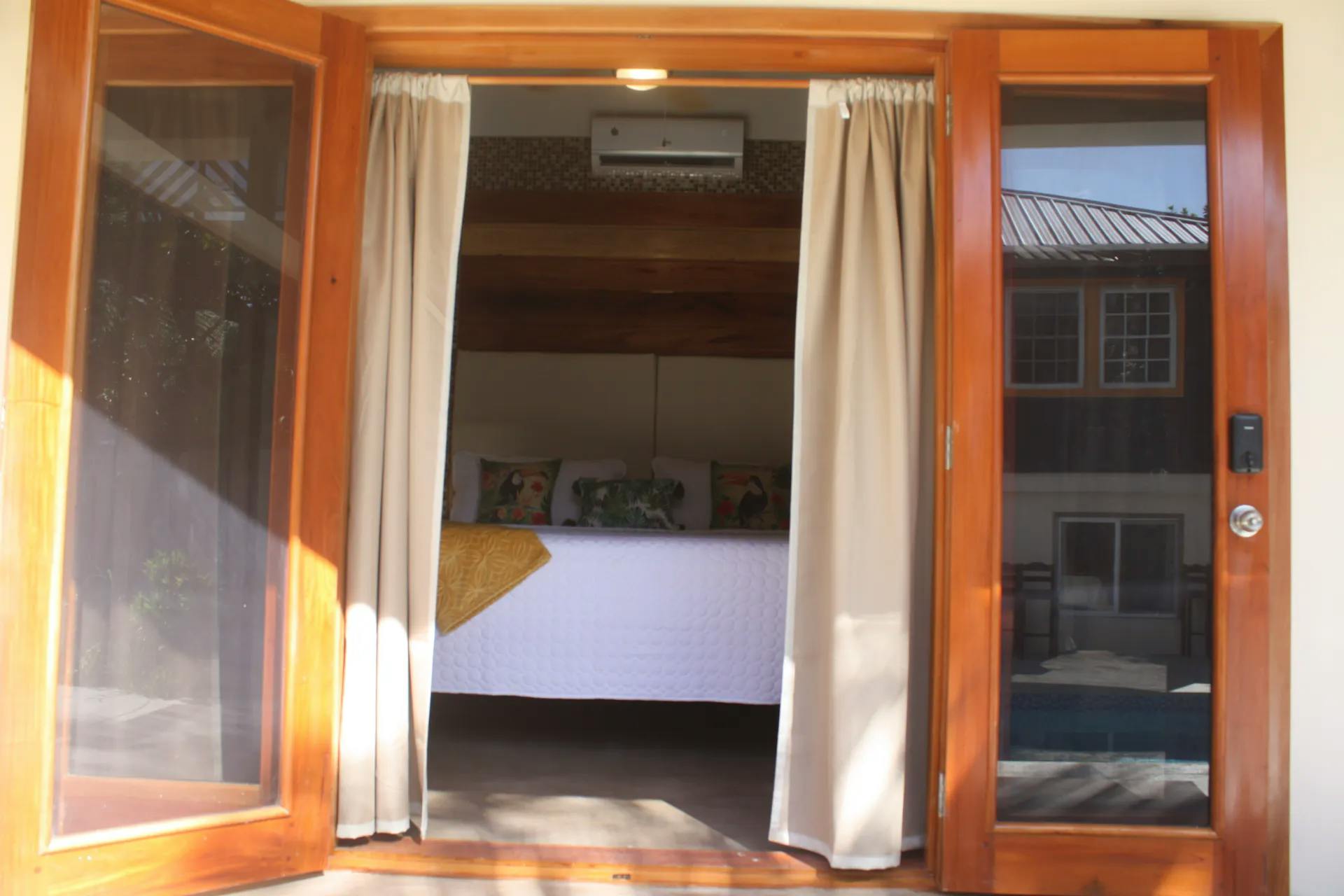 Looking through wooden draped french doors into bedroom with white bed cover and decorative throw pillows with toucans and tropical foliage