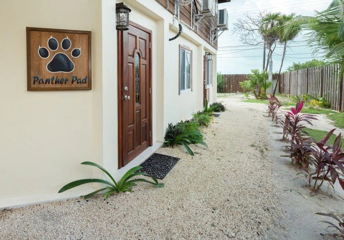 Stone walkway with tropical foliage leading to exterior dark wooden door and animal paw print sign with the words "Panther Pad"