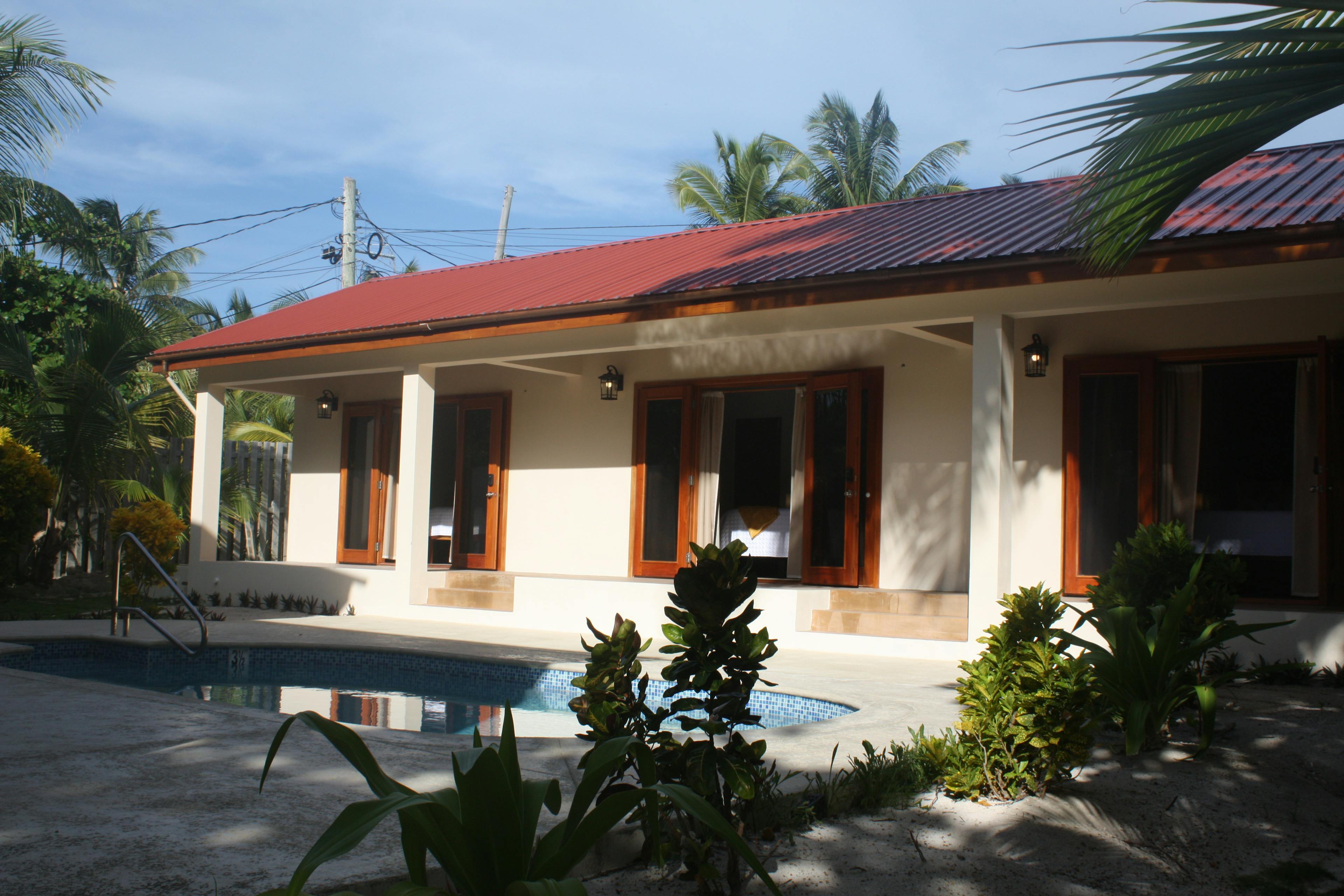 Pool in the foreground with steps leading to long white building with red roof and three sets of wooden french doors